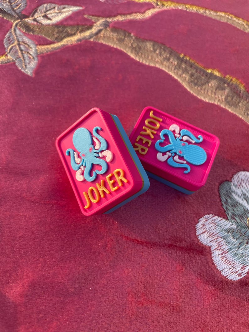 Puede incluir: Dos fichas de juego rectangulares con una superficie superior de color rosa brillante, una base azul claro y un gr&aacute;fico de pulpo azul claro en relieve. La palabra JOKER est&aacute; grabada en letras amarillas sobre la superficie rosa de cada ficha.