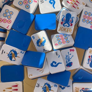 May include: A collection of white and blue Mahjong tiles. The tiles feature various symbols, numbers, and the word "JOKER". The tiles are scattered, with some blue tiles interspersed among them. The image is taken from a high angle.