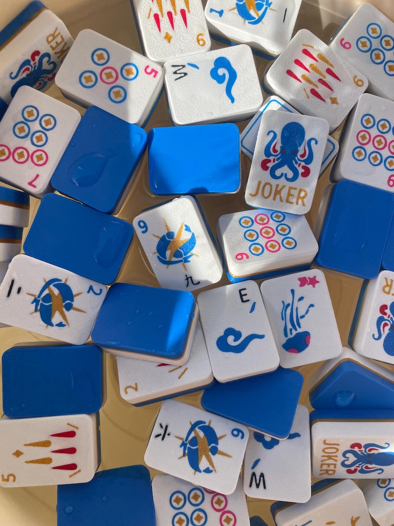May include: A collection of white and blue Mahjong tiles. The tiles feature various symbols, numbers, and the word "JOKER". The tiles are scattered, with some blue tiles interspersed among them. The image is taken from a high angle.