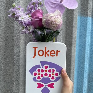 May include: A white rectangular vase with the word "Joker" in orange lettering and a purple floral bouquet illustration. The vase holds a variety of purple and pink flowers, including roses and anthurium.