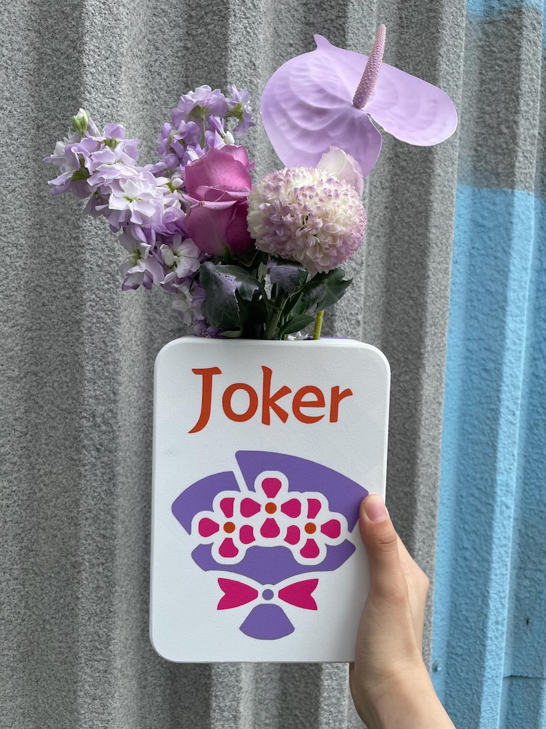 May include: A white rectangular vase with the word "Joker" in orange lettering and a purple floral bouquet illustration. The vase holds a variety of purple and pink flowers, including roses and anthurium.