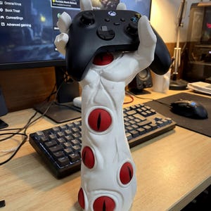 May include: A white, stylized hand-shaped gaming controller holder with red eye accents. The holder is designed to cradle a black Xbox controller. A unique and decorative accessory for gamers.
