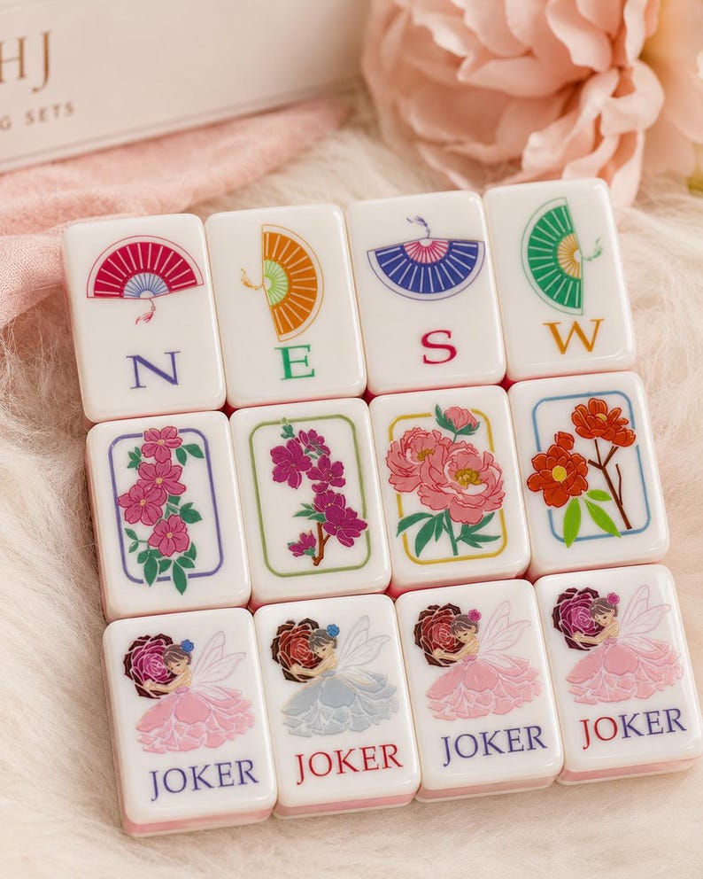 May include: A set of white Mahjong tiles with colorful illustrations. The tiles feature images of fans, flowers, and fairies. Some tiles have the letters N, E, S, and W. The word "JOKER" is printed on several tiles.