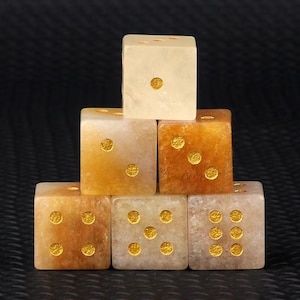 May include: Six dice stacked in a pyramid formation. The dice are a mix of light tan and amber colors, with gold-colored dots indicating the numbers. The dice are made of a translucent material, and the background is black.
