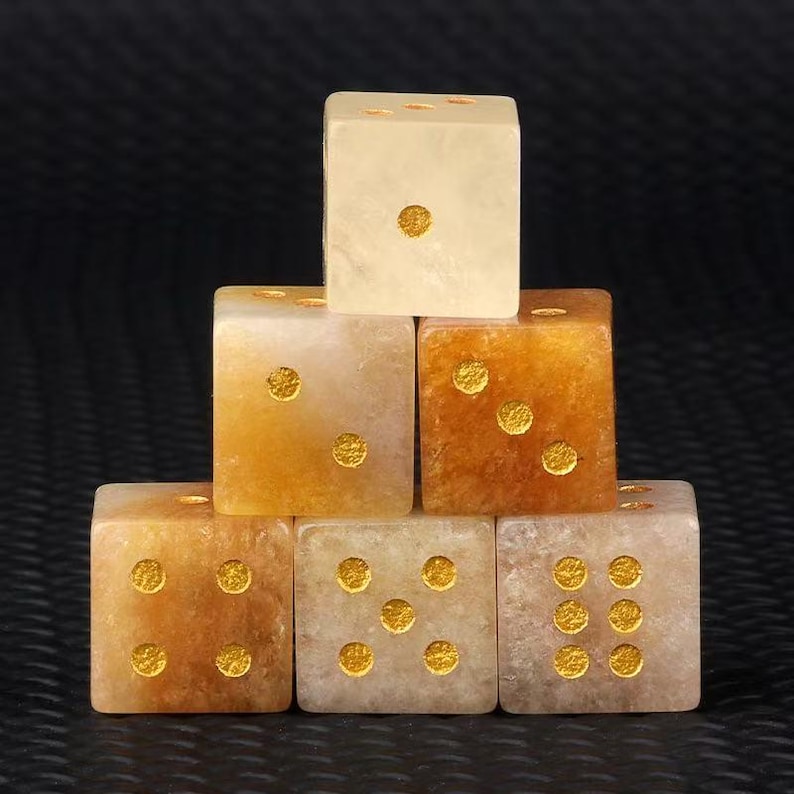 May include: Six dice stacked in a pyramid formation. The dice are a mix of light tan and amber colors, with gold-colored dots indicating the numbers. The dice are made of a translucent material, and the background is black.