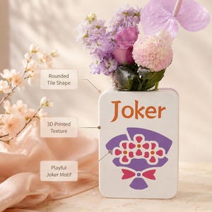 May include: A white ceramic vase with the word "Joker" in orange, holding a bouquet of purple, pink, and white flowers. The vase has a 3D printed texture and a playful Joker motif with a bouquet design.