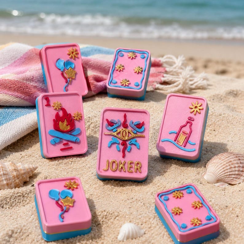 Puede incluir: Cartas de juego rosas y azules con varios dise&ntilde;os, incluyendo una hoguera y una botella, dispuestas sobre una playa de arena. Una carta dice "JOKER" en letras doradas. Las cartas tienen forma rectangular.