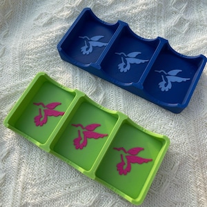 Puede incluir: Dos bandejas decorativas, una azul y otra verde, cada una con tres compartimentos. La bandeja azul presenta un dise&ntilde;o de colibr&iacute; blanco, mientras que la bandeja verde tiene un dise&ntilde;o de colibr&iacute; rosa. Las bandejas son de pl&aacute;stico.