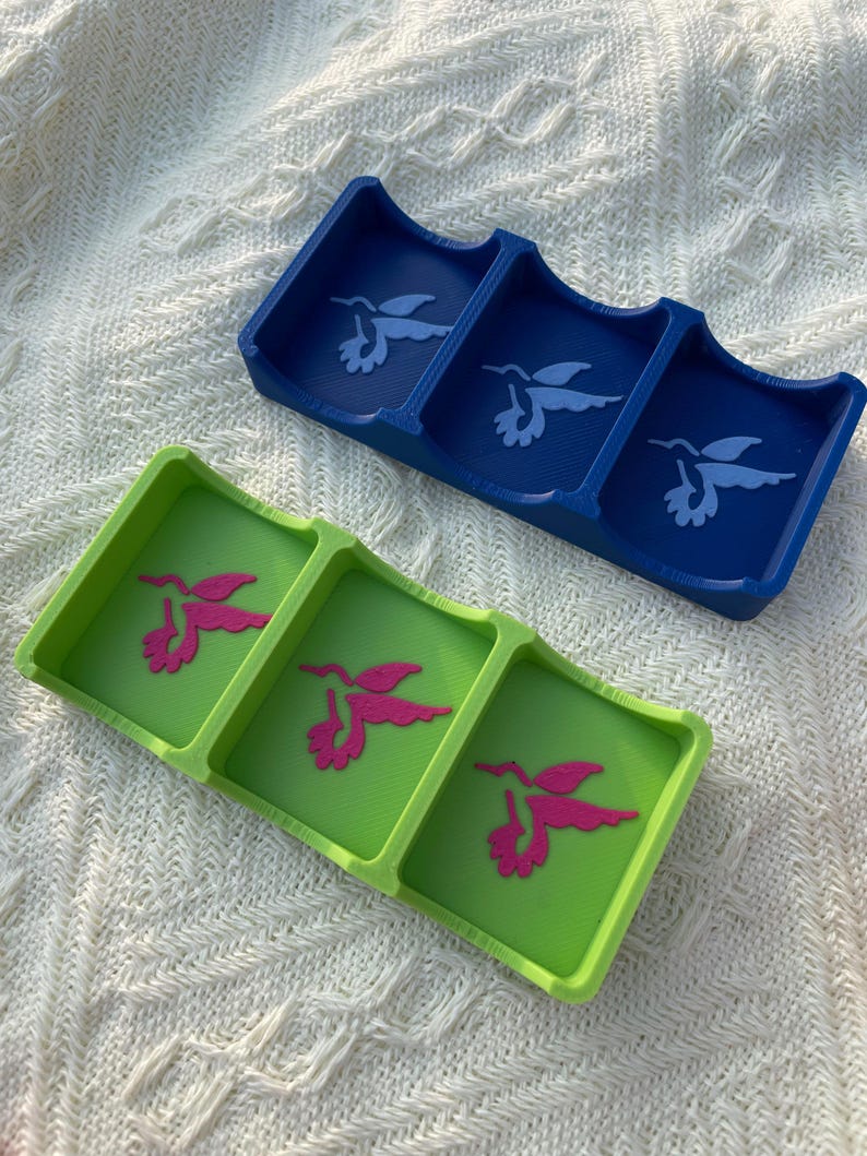 Puede incluir: Dos bandejas decorativas, una azul y otra verde, cada una con tres compartimentos. La bandeja azul presenta un dise&ntilde;o de colibr&iacute; blanco, mientras que la bandeja verde tiene un dise&ntilde;o de colibr&iacute; rosa. Las bandejas son de pl&aacute;stico.
