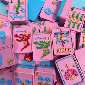 Puede incluir: Un conjunto de fichas de Mahjong rosas con detalles azules y dorados. Las fichas presentan varios s&iacute;mbolos, incluyendo dragones, flores y n&uacute;meros. Algunas fichas tienen un borde azul, otras un borde azul y dorado. El fondo es azul claro.