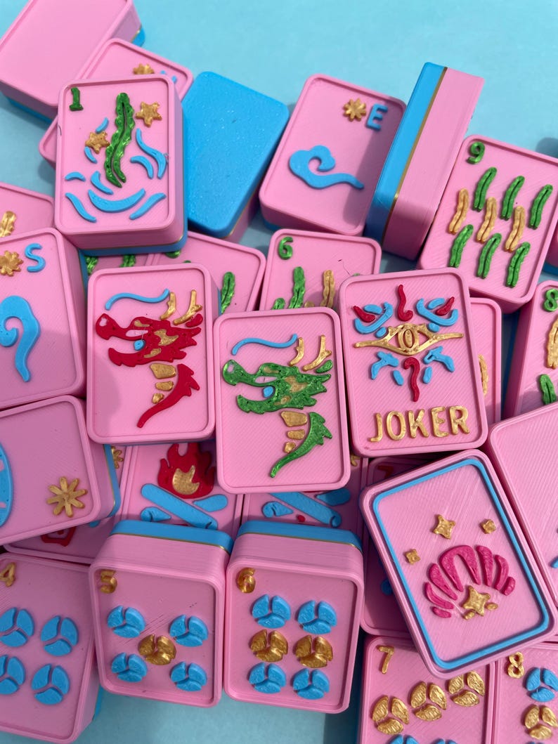 Puede incluir: Un conjunto de fichas de Mahjong rosas con detalles azules y dorados. Las fichas presentan varios s&iacute;mbolos, incluyendo dragones, flores y n&uacute;meros. Algunas fichas tienen un borde azul, otras un borde azul y dorado. El fondo es azul claro.