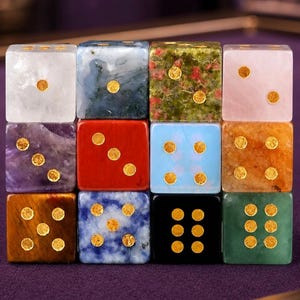 May include: A collection of twelve gemstone dice in various colors and patterns. Each die features gold-colored dots indicating the numbers. The dice are arranged in a grid, showcasing the diverse textures and hues of the gemstones, including amethyst, red jasper, and green jade.