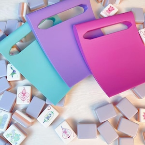 May include: Three solid-colored plastic tile racks in light blue, purple, and hot pink, each featuring a rectangular handle cutout, arranged over a scattered collection of rectangular mahjong tiles with various colorful printed designs.