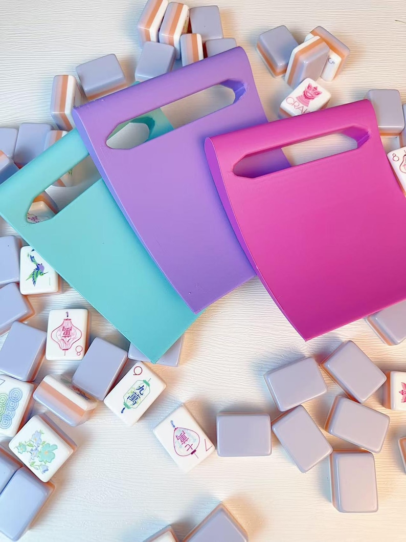 May include: Three solid-colored plastic tile racks in light blue, purple, and hot pink, each featuring a rectangular handle cutout, arranged over a scattered collection of rectangular mahjong tiles with various colorful printed designs.