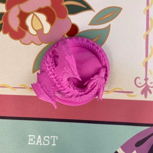 May include: A bright magenta 3D printed dragon figurine coiled in a circular shape. The dragon features intricate scale textures, visible wings, and a detailed head. The surface of the figurine shows fine horizontal layer lines from the printing process.