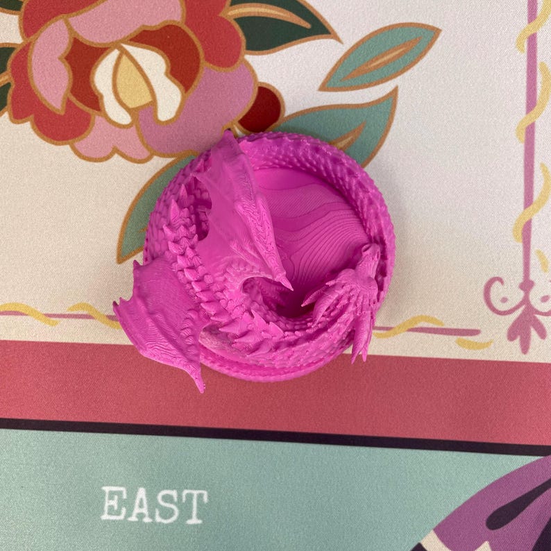 May include: A bright magenta 3D printed dragon figurine coiled in a circular shape. The dragon features intricate scale textures, visible wings, and a detailed head. The surface of the figurine shows fine horizontal layer lines from the printing process.