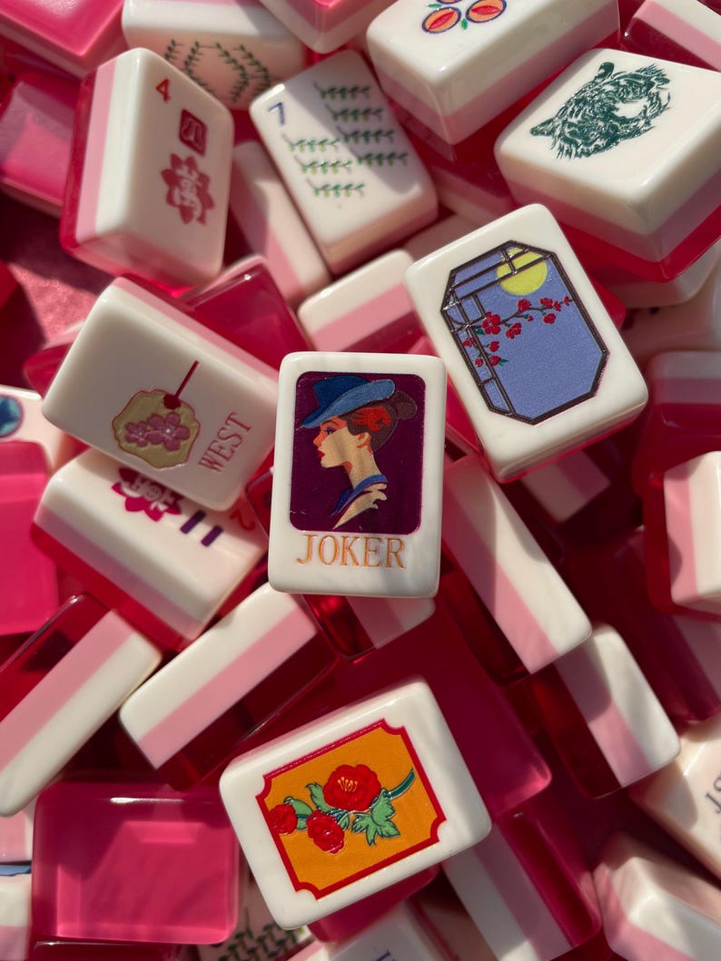 May include: A collection of white and pink Mahjong tiles with various illustrations and numbers. One tile features a woman in a hat with the word "JOKER". Other tiles display floral designs, a cherry, and a tiger.