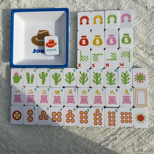 Może przedstawiać: Kompletny zestaw do gry w Mahjong z białymi kafelkami z r&oacute;żnymi symbolami i liczbami. Zawiera tacę z niebieską obw&oacute;dką z grafiką kapelusza kowbojskiego i słowem "JOKER". Kafelki przedstawiają podkowy, worki z pieniędzmi i symbole kierunk&oacute;w.