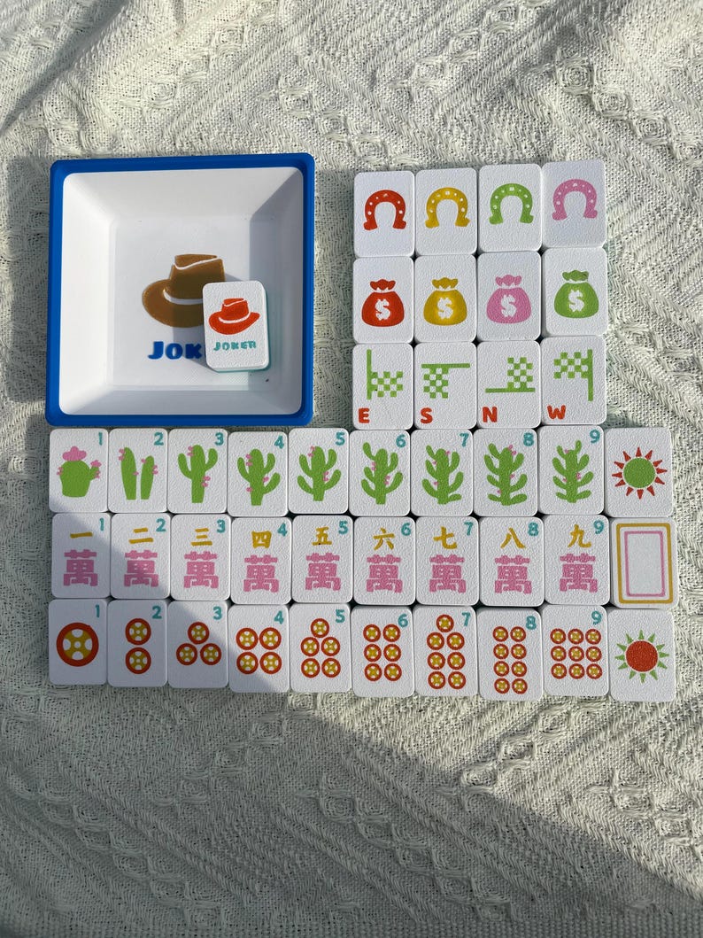Może przedstawiać: Kompletny zestaw do gry w Mahjong z białymi kafelkami z r&oacute;żnymi symbolami i liczbami. Zawiera tacę z niebieską obw&oacute;dką z grafiką kapelusza kowbojskiego i słowem "JOKER". Kafelki przedstawiają podkowy, worki z pieniędzmi i symbole kierunk&oacute;w.