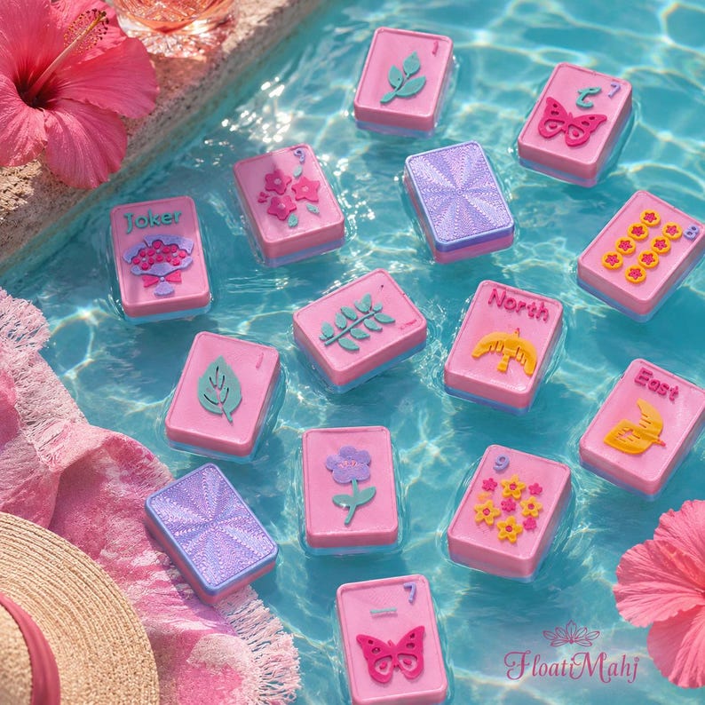 May include: Pink and blue Mahjong tiles floating in water. The tiles feature various designs, including flowers, butterflies, and the words "Joker", "North", and "East". A straw hat and pink flowers are in the background.