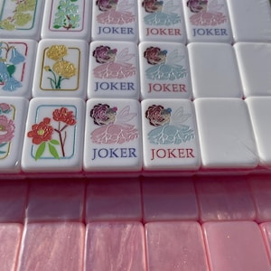 May include: A set of white Mahjong tiles with colorful floral designs and the word "JOKER" printed on some. The tiles feature various flower illustrations in shades of pink, yellow, and blue. The bottom row shows pink tiles.