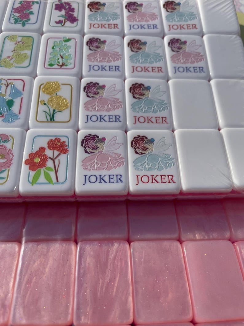May include: A set of white Mahjong tiles with colorful floral designs and the word "JOKER" printed on some. The tiles feature various flower illustrations in shades of pink, yellow, and blue. The bottom row shows pink tiles.