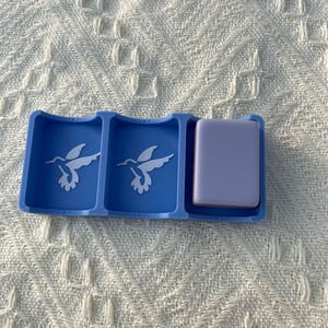 Puede incluir: Un organizador azul con tres compartimentos. Dos compartimentos presentan un dise&ntilde;o de colibr&iacute; blanco. El tercer compartimento contiene un objeto rectangular de color lavanda. El organizador est&aacute; sobre una superficie texturizada blanquecina.