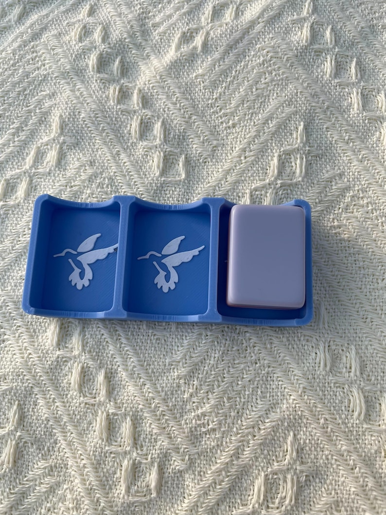 Puede incluir: Un organizador azul con tres compartimentos. Dos compartimentos presentan un dise&ntilde;o de colibr&iacute; blanco. El tercer compartimento contiene un objeto rectangular de color lavanda. El organizador est&aacute; sobre una superficie texturizada blanquecina.