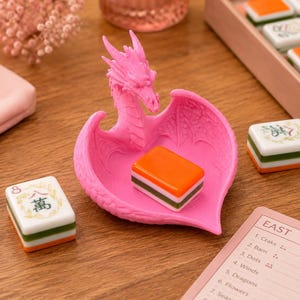 Pu&ograve; includere: Un vassoio a forma di cuore di colore rosa brillante con una figura di drago, ali testurizzate e una testa di drago dettagliata. Una singola tessera del Mahjong arancione, bianca e verde riposa nel vassoio, circondata da altre tessere e un segnapunti.