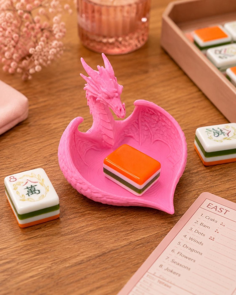 Pu&ograve; includere: Un vassoio a forma di cuore di colore rosa brillante con una figura di drago, ali testurizzate e una testa di drago dettagliata. Una singola tessera del Mahjong arancione, bianca e verde riposa nel vassoio, circondata da altre tessere e un segnapunti.