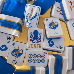 Könnte beinhalten: Nahaufnahme von Mahjong-Steinen in Blau, Weiß und Gold. Die Steine zeigen verschiedene Symbole und Zahlen, darunter einen Oktopus, Seetang und das Wort "JOKER". Die Steine sind eng aneinandergereiht und bilden ein ansprechendes Muster.