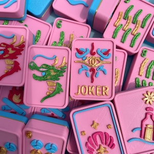 May include: A collection of pink Mahjong tiles with blue and gold accents. The tiles feature various embossed designs, including dragons, clouds, and the word "JOKER". The game pieces are arranged in a close-up shot.