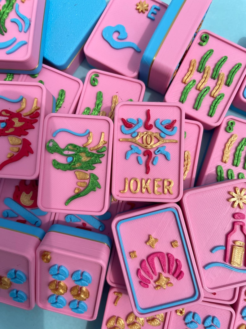Puede incluir: Un conjunto de fichas de Mahjong rosas con detalles azules y dorados. Las fichas presentan varios dise&ntilde;os en relieve, incluyendo dragones, nubes y la palabra "JOKER". Las piezas del juego est&aacute;n dispuestas en un primer plano.