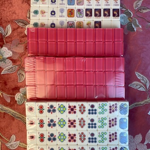 May include: A new Mahjong set with white tiles featuring various symbols and numbers, including "EAST", "WEST", "SOUTH", "NORTH", and "JOKER". The set includes pink tile racks, all packaged in clear plastic.