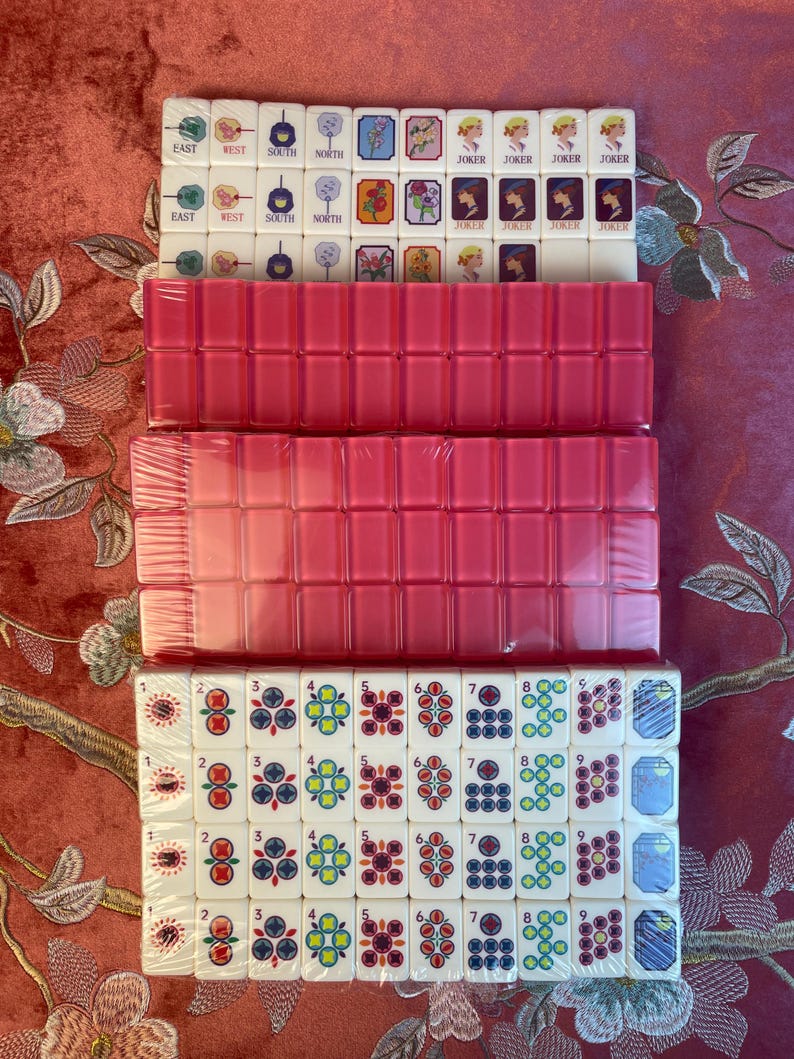 May include: A new Mahjong set with white tiles featuring various symbols and numbers, including "EAST", "WEST", "SOUTH", "NORTH", and "JOKER". The set includes pink tile racks, all packaged in clear plastic.