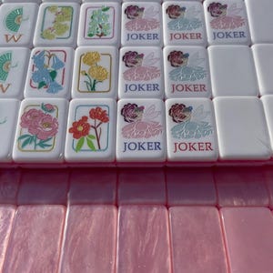 May include: A collection of white Mahjong tiles with colorful floral designs and the word "JOKER" on some. The tiles are arranged on a pink surface, creating a visually appealing pattern. The floral designs include pink, yellow, and blue flowers.