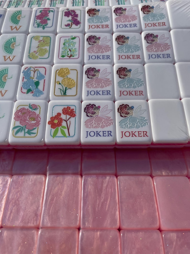 May include: A collection of white Mahjong tiles with colorful floral designs and the word "JOKER" on some. The tiles are arranged on a pink surface, creating a visually appealing pattern. The floral designs include pink, yellow, and blue flowers.