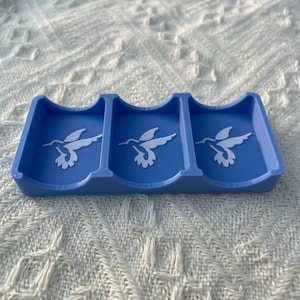 Puede incluir: Un tarjetero azul con tres secciones, cada una con un dise&ntilde;o de colibr&iacute; blanco. El soporte est&aacute; hecho de un material s&oacute;lido y est&aacute; dise&ntilde;ado para guardar tarjetas. El fondo es una superficie blanca texturizada.