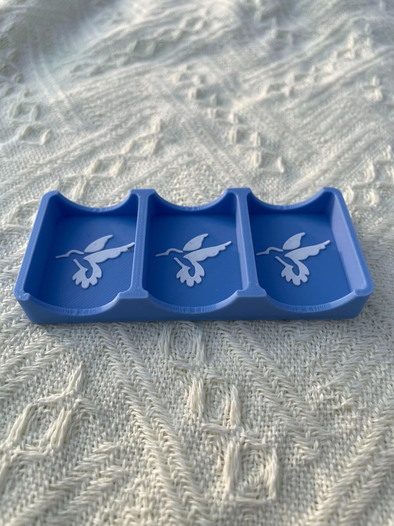 Puede incluir: Un tarjetero azul con tres secciones, cada una con un dise&ntilde;o de colibr&iacute; blanco. El soporte est&aacute; hecho de un material s&oacute;lido y est&aacute; dise&ntilde;ado para guardar tarjetas. El fondo es una superficie blanca texturizada.