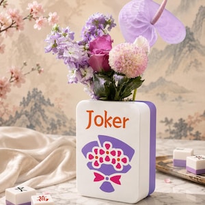 May include: A white and purple ceramic vase with the word "Joker" in orange, holding a bouquet of purple, pink, and white flowers. The vase has a floral design. Several Mahjong tiles are in the foreground.
