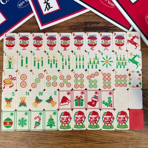 May include: A set of off-white mahjong tiles featuring festive holiday illustrations including Christmas trees, reindeer, Santa hats, stockings, gifts, and candles. The tiles are arranged in a grid on a wooden surface with game instruction cards.