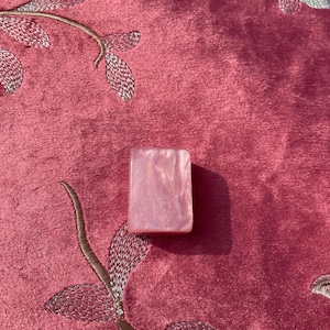 May include: A rectangular, pink, iridescent stone. The stone has a pearlescent sheen and is set against a pink, embroidered fabric background. The fabric has a floral design.