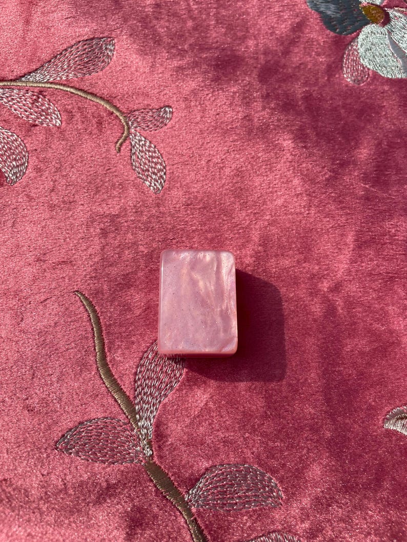 May include: A rectangular, pink, iridescent stone. The stone has a pearlescent sheen and is set against a pink, embroidered fabric background. The fabric has a floral design.