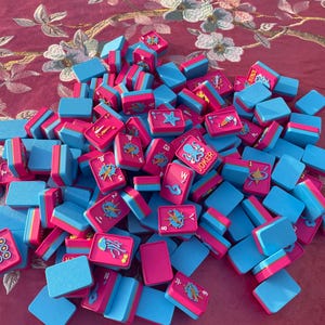 Puede incluir: Una colecci&oacute;n de fichas de mahjong rectangulares con un esquema de colores rosa vibrante y azul brillante. Las fichas muestran varias ilustraciones coloridas, incluyendo criaturas marinas como un pulpo etiquetado como JOKER, una medusa y una estrella de mar.