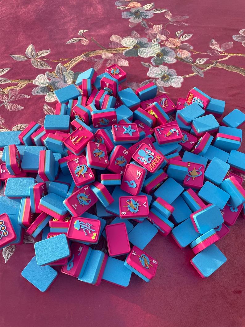 Puede incluir: Una colecci&oacute;n de fichas de mahjong rectangulares con un esquema de colores rosa vibrante y azul brillante. Las fichas muestran varias ilustraciones coloridas, incluyendo criaturas marinas como un pulpo etiquetado como JOKER, una medusa y una estrella de mar.