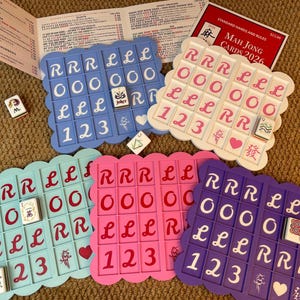 Könnte beinhalten: Ein farbenfrohes Mahjong-Set mit vier wellenförmigen Spielbrettern in Blau, Rosa, Türkis und Lila. Jedes Brett hat ein Raster aus Spielsteinen mit Zahlen und Symbolen. Das Set enthält Spielsteine, ein Regelbuch und eine Box mit der Aufschrift "Mah Jong Cards 2026".