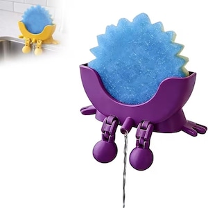 May include: A purple crab-shaped sponge holder with a blue sponge. The holder has articulated legs and a spout for water drainage. A yellow version of the holder is visible in the background. The sponge has a starburst shape.