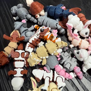 May include: A collection of articulated animal toys in various colors. The toys include a gray koala, a brown turtle, a blue dragon, a white rabbit with pink accents, and a tiger-striped gecko. Other animals include a raccoon and a mouse.