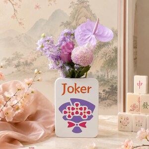 May include: A white ceramic vase with the word "Joker" in orange lettering and a purple floral design. The vase holds a bouquet of purple and pink flowers. Mahjong tiles and a box are in the background.