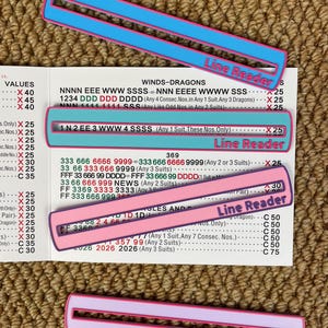 Op de afbeelding: Vier kleurrijke Line Reader-geleiders in blauw en roze met witte tekst, rustend op een bedrukte spelkaart. De geleiders hebben een rechthoekige vorm met een uitsparing in het midden. De spelkaart heeft zwarte tekst.